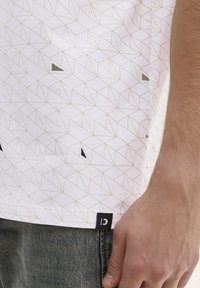 Camisa blanca de manga corta con un patrón geométrico en gris claro. Presenta acentos triangulares negros y una pequeña etiqueta negra en el dobladillo.