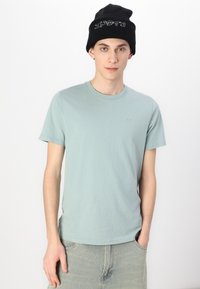 Ανοιχτό μπλε κοντομάνικο μπλουζάκι από μαλακό ύφασμα, σε συνδυασμό με ανοιχτόχρωμες τζιν παντελόνες. Μαύρο πλεκτό σκουφάκι με λευκές λεπτομέρειες κειμένου.