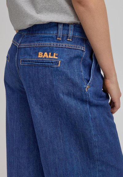 Sinibug na jeans na asul na denim na may kahel na tahi. Kasama sa mga tampok ang mga likod na bulsa at nakaburdang "BALL" na tatak sa kanang bahagi.