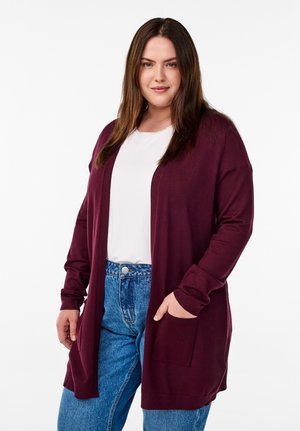 Femme portant une chemise blanche, un jean bleu et un long cardigan bordeaux avec des poches, debout sur un fond blanc uni.