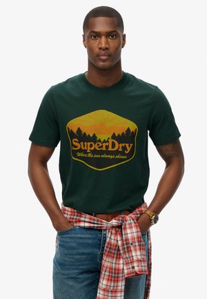 Groene katoenen T-shirt met een vintage-stijl logo met een zon en boom graphic. Het model draagt een geruit overhemd om de taille geknoopt.