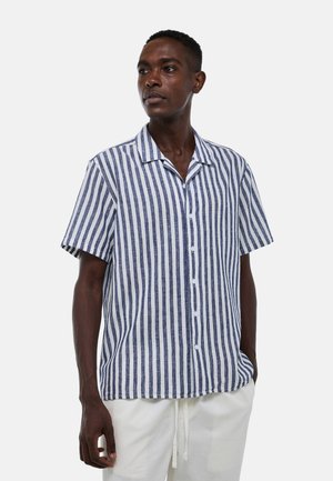 Homme portant une chemise à manches courtes à rayures verticales bleues et blanches avec des boutons blancs et un pantalon blanc, debout devant un fond uni.