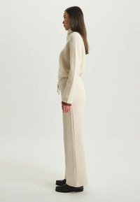 Pull en maille côtelée beige à col roulé associé à un pantalon ample assorti. Les deux vêtements présentent une texture douce et un détail de cordon de serrage. Chaussures noires.