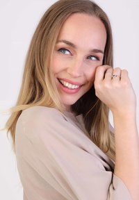 Eine Frau mit langen, welligen blonden Haaren trägt ein beiges Oberteil. Sie lächelt, berührt ihr Gesicht und präsentiert einen silbernen Ring mit einem blauen Edelstein.