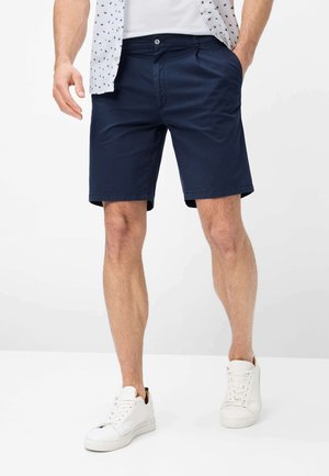 Uomo che indossa pantaloncini blu navy, scarpe da ginnastica bianche, una camicia bianca e una camicia a fantasia parzialmente infilata, con una mano in tasca.