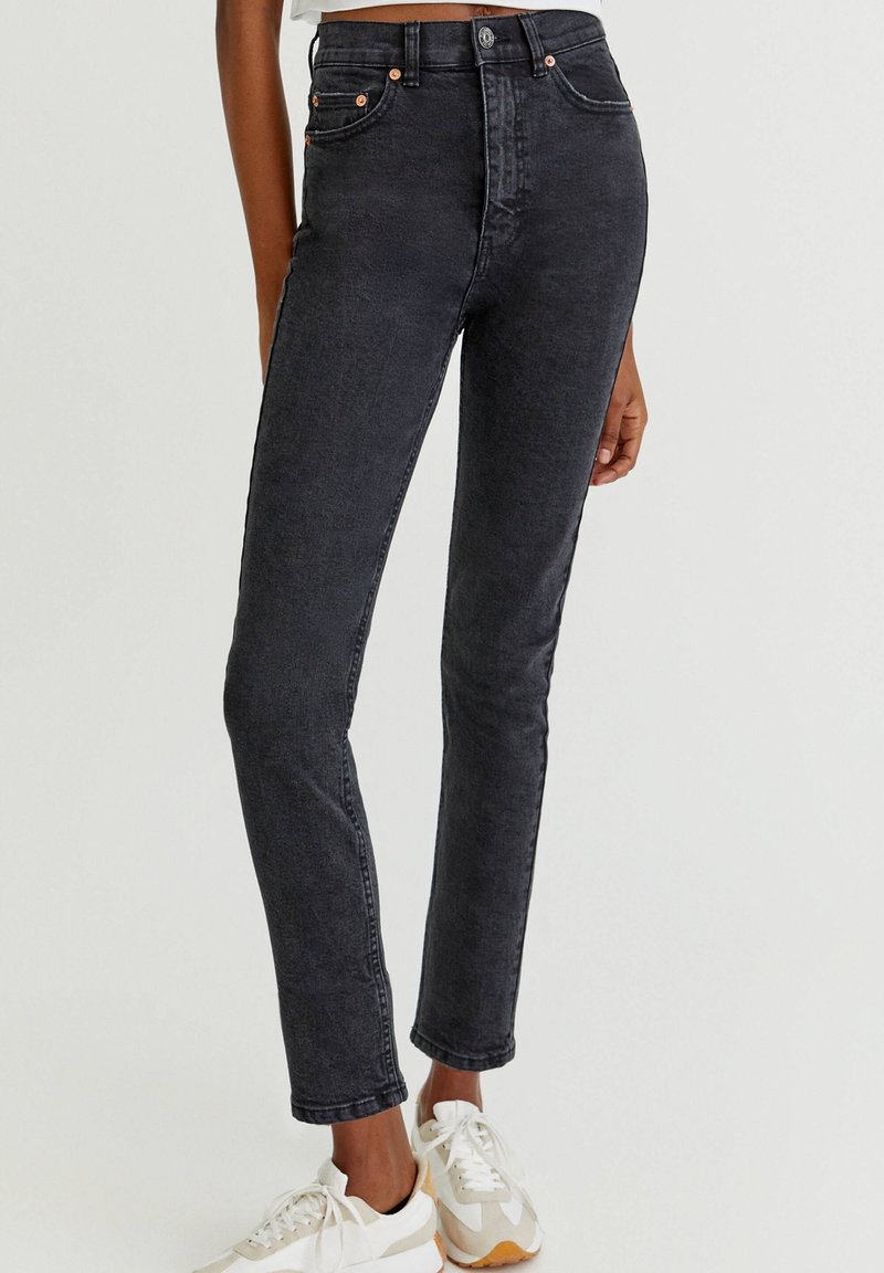 Jean noir taille haute ajusté, porté avec des baskets blanches et beiges, montré de la taille aux chevilles sur une personne debout.