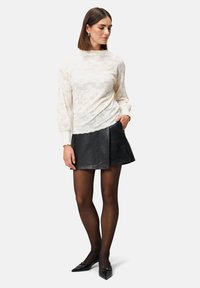 Cremfarbener, strukturierter Pullover mit langen Ärmeln, kombiniert mit einem schwarzen Leder-Minikleid. Das Model trägt transparente schwarze Strumpfhosen und spitze Schuhe.
