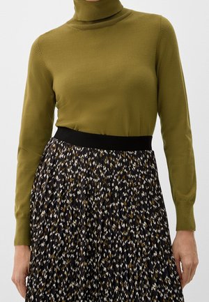 Femme portant un pull à col roulé vert olive rentré dans une jupe plissée taille haute à motifs noirs, blancs et bruns.