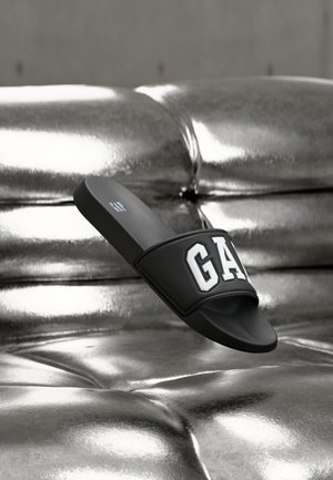 Sandale noire à enfiler avec le texte blanc "GAP" sur un fond de canapé matelassé argenté métallique.