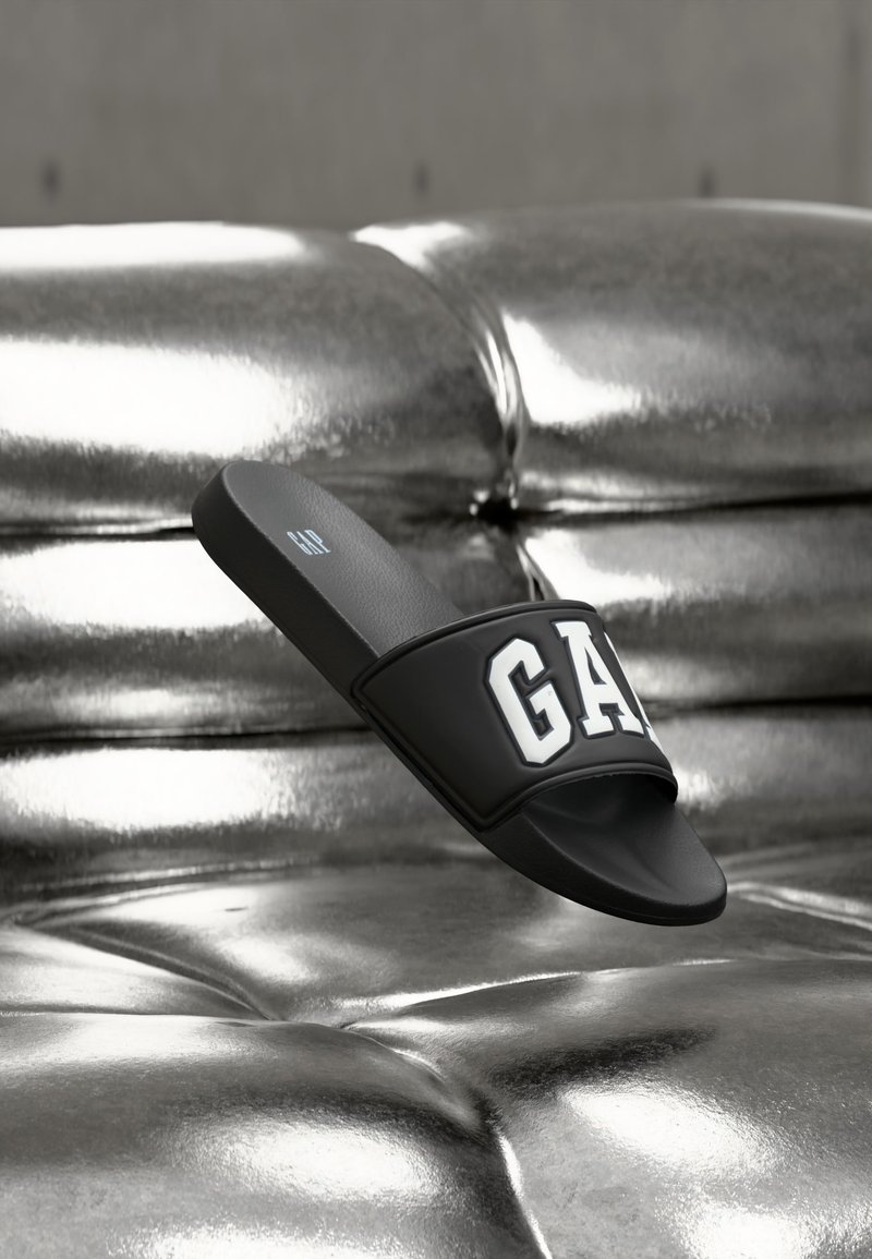 Sandale noire à enfiler avec le texte blanc "GAP" sur un fond de canapé matelassé argenté métallique.