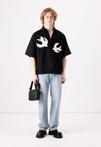 Černá košile s krátkým rukávem s bílými motivy ptáků, zkombinována se světle modrými džíny a černými robustními botami. Drží černou kabelku.