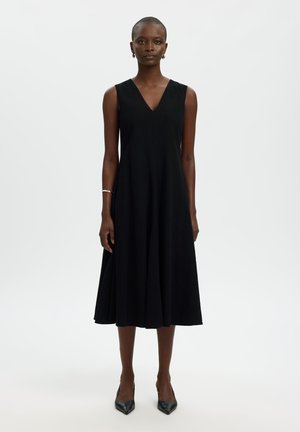 Kvinne som står iført en ermeløs svart midi-kjole med V-hals, svarte spisse hæler, sølvarmbånd og hoopearrings på en hvit bakgrunn.