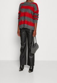 Person med en röd- och grårandig stickad tröja, svarta läderbyxor, svarta klackstövlar med silvertippar, som håller en grå handväska.