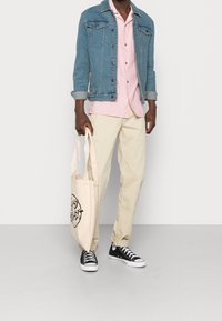 Uomo con camicia rosa chiaro, giacca di jeans blu, pantaloni crema e sneakers nere alte, che tiene una borsa a tracolla beige con stampa nera.