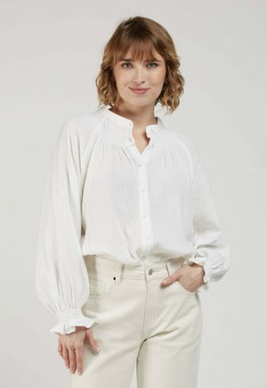 Femme aux cheveux courts et ondulés portant un chemisier blanc boutonné avec des manches froncées et un pantalon crème taille haute, debout avec une main dans la poche.