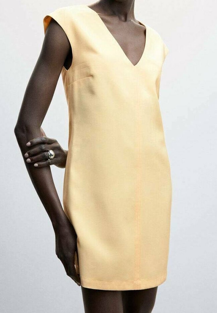 Personne à la peau foncée portant une robe à manches courtes, jaune pâle, en V avec une silhouette ajustée, main posée sur la hanche, bague argentée au doigt.