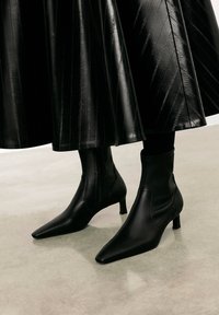 Botines negros de cuero suave con punteras afiladas y tacones bajos. El material tiene un sutil brillo, complementando una falda negra plisada.