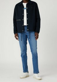 Dark navy jacket with a zippered pocket, silver snap buttons, and a logo patch. Paired with light blue jeans and white sneakers.