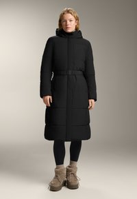 Geacă puffer neagră cu talie accentuată, mâneci lungi și guler înalt. Îmbogățită cu o textură matlasată și glugă. Asortată cu cizme căptușite cu blană maronie.