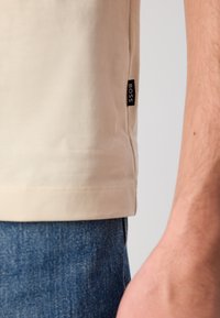 Beige shirt hem with black BOSS label worn over blue denim jeans, showing part of a person's arm against a plain background.