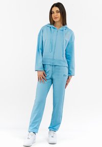 Felpa zip con cappuccio azzurra chiara a maniche lunghe, abbinata a pantaloni della tuta coordinati. Realizzata in tessuto morbido, presenta un logo sul petto. Sneakers bianche.