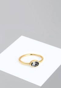 Anello d'oro con una pietra ovale grigia montata su una semplice fascia, posato su una superficie bianca. Finitura liscia, design minimalista.