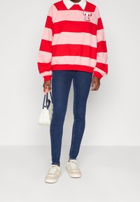 Sudadera a rayas rojas y rosas con cuello blanco, jeans skinny azules y zapatillas beige. Lleva un bolso pequeño en la mano, con un corte relajado y puños acanalados.