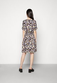 Vestido floral em tecido escuro, com mangas curtas, cintura ajustada e saia rodada. Detalhes de flores roxas, amarelas e brancas. Sapatos de salto bloco pretos.