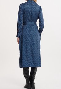 Długa niebieska sukienka z gładkiego materiału, z wzorem diamentów, z paskiem w talii i mankietami na guziki. Noszona z czarnymi botkami do kolan.