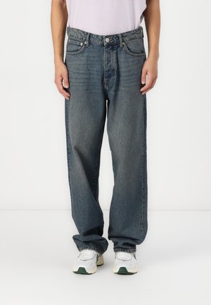 Baggy blue denim jeans with a faded finish, five-pocket design, and a button closure. Worn with white sneakers.