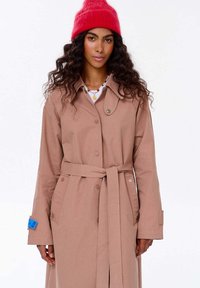 Kobieta z długimi kręconymi włosami, ubrana w beżowy płaszcz z paskiem, nałożone naszyjniki i jasnoczerwony, dziany kapelusz, stojąca na tle białym.