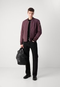 Burgundy bomber jacket with ribbed cuffs, zip pocket detail, black t-shirt, black pants, holding a black duffel bag, chunky black boots.