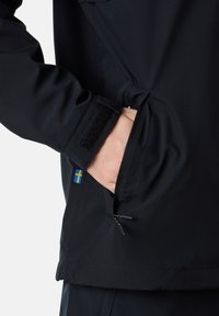 Svart vattentät jacka som visar en sidficka med dragkedja, kardborrestängning vid ärmarna och en liten svensk flagga som patch på ärmen.