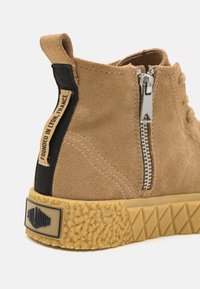 Baskets en suède beige avec une semelle en caoutchouc texturé. Présentent une fermeture éclair sur le côté et un tirant en tissu noir avec le texte "Fondé à Lyon, France".