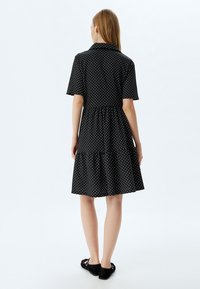 Vestido negro de manga corta con lunares blancos, diseño con cuello, cintura elástica y falda con volantes. Lo lleva puesto con zapatos de ante negros.
