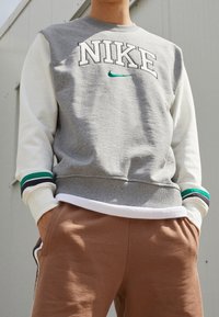 Person som bär en grå Nike-tröja med vita ärmar och bruna byxor med händerna i fickorna, stående utomhus.