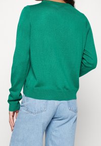 Grüner Strickpullover mit langen Ärmeln und rundem Ausschnitt, ausgestattet mit einem gerippten Saum und Bündchen. Kombiniert mit hellblauen Jeans mit hoher Taille.