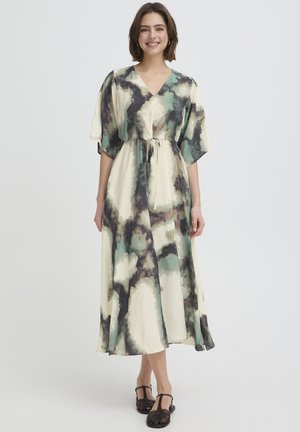 Žena v dlhých, voľných šatách so zelenými, čiernymi a krémovými abstraktnými vzormi, stojaca a usmievajúca sa na jednoduchom pozadí.