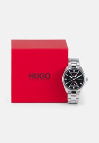 Edelstahl-Uhr mit schwarzem Zifferblatt, silbernen Akzenten und einem Metallarmband, neben einer roten Box mit dem "HUGO"-Logo.
