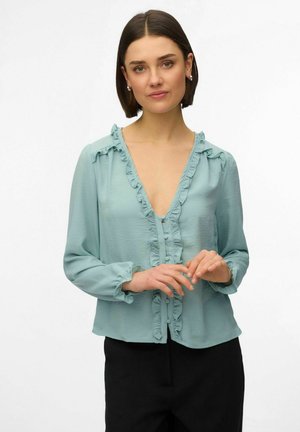 Femme aux cheveux bruns courts portant un chemisier bleu clair à volants avec des boutons, et un pantalon noir, posant sur un fond blanc.