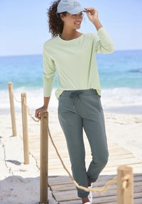 Hellgrünes langärmliges Shirt, graue Jogginghose mit Kordelzug und eine hellgraue Mütze. Sand und Ozean im Hintergrund mit einem Holzweg.