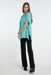 Turkio žalia megzta pončo su kaklo apykakle, su kutais išilgai kraštų, derinama su juodomis tiesiomis kelnėmis ir juodais batais.