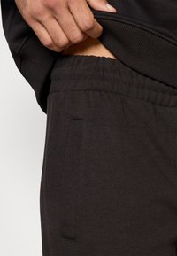 Hnedé bavlnené tepláky s elastickým pásom a bočnými vreckami, zobrazené vedľa ruky, ktorá drží pás. Štruktúra vyzerá mäkko.