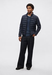 Marineblå quiltet puffervest over en ribstrikket sweater, kombineret med mørk denim jeans med vide ben og sorte sko. Enkel, funktionel design.