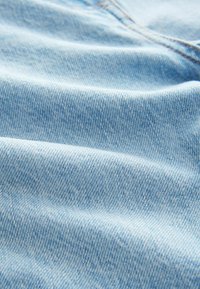 Tejido de denim azul claro con una textura suave y un tejido de sarga visible. Presenta variaciones sutiles en el tono y detalles de costura finos.