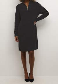 Femme portant une robe en maille noire longueur genou avec un col à fermeture éclair dorée et des chaussures à talons noires, debout avec une main sur la hanche.