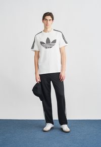 Λευκό μπλουζάκι με μαύρο λογότυπο, μαύρο αθλητικό παντελόνι με λευκές πλευρικές ρίγες και λευκά αθλητικά παπούτσια. Το μοντέλο κρατάει μαύρο καπέλο.