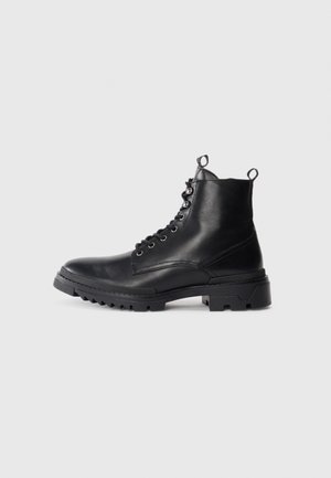 Botas de tobillo de cuero negro con una suela robusta, diseño con cordones y dos lengüetas en el eje para facilitar su uso. Textura suave y acentos mínimos.