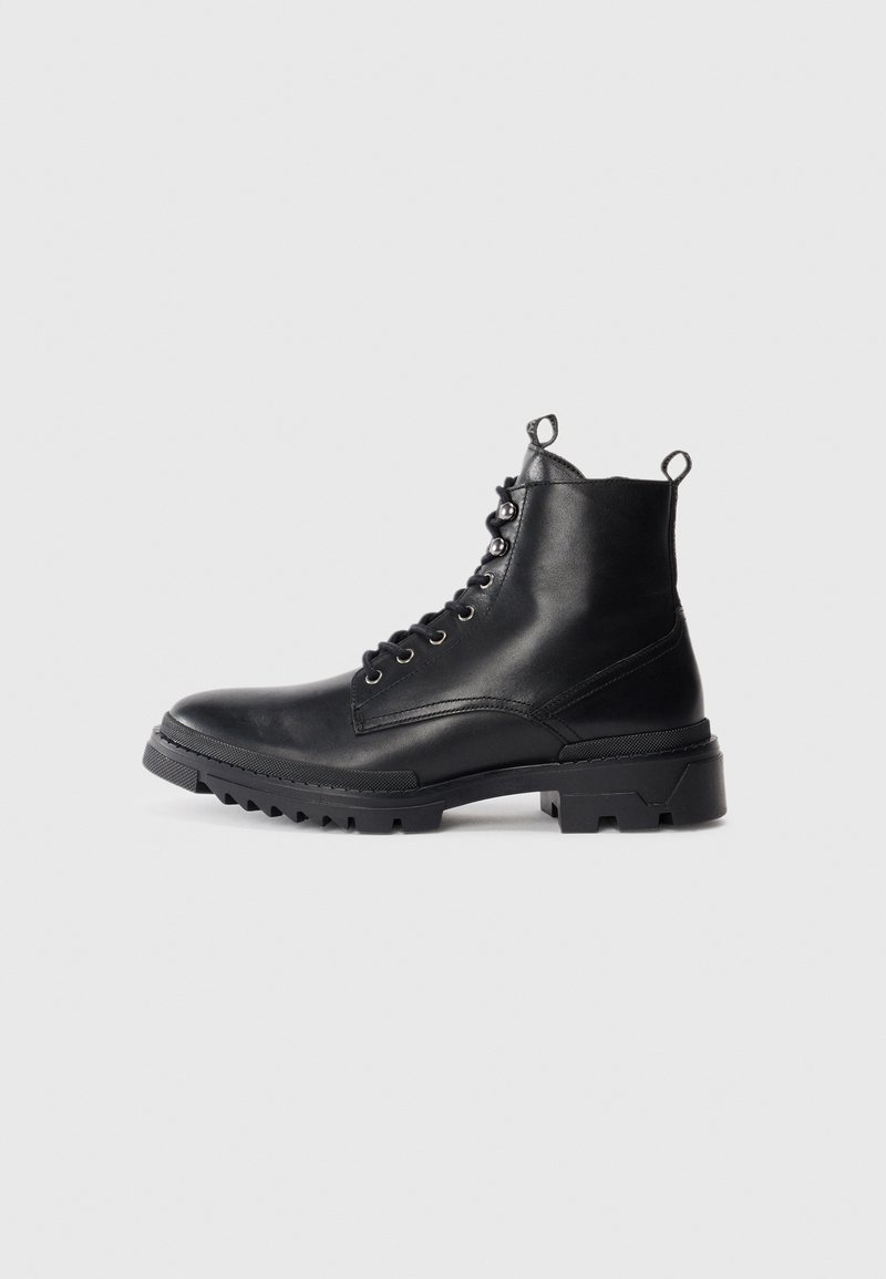 Botas de tobillo de cuero negro con una suela robusta, diseño con cordones y dos lengüetas en el eje para facilitar su uso. Textura suave y acentos mínimos.