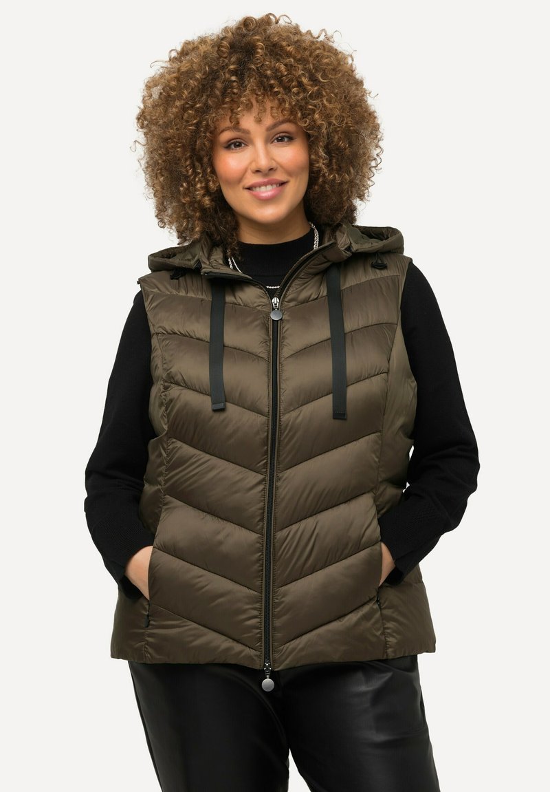 Olivově zelená prošívaná vesta s kapucí, s žebrovaným prošíváním, předním zipem a černými nastavitelnými popruhy. Nositá přes černý svetr.
